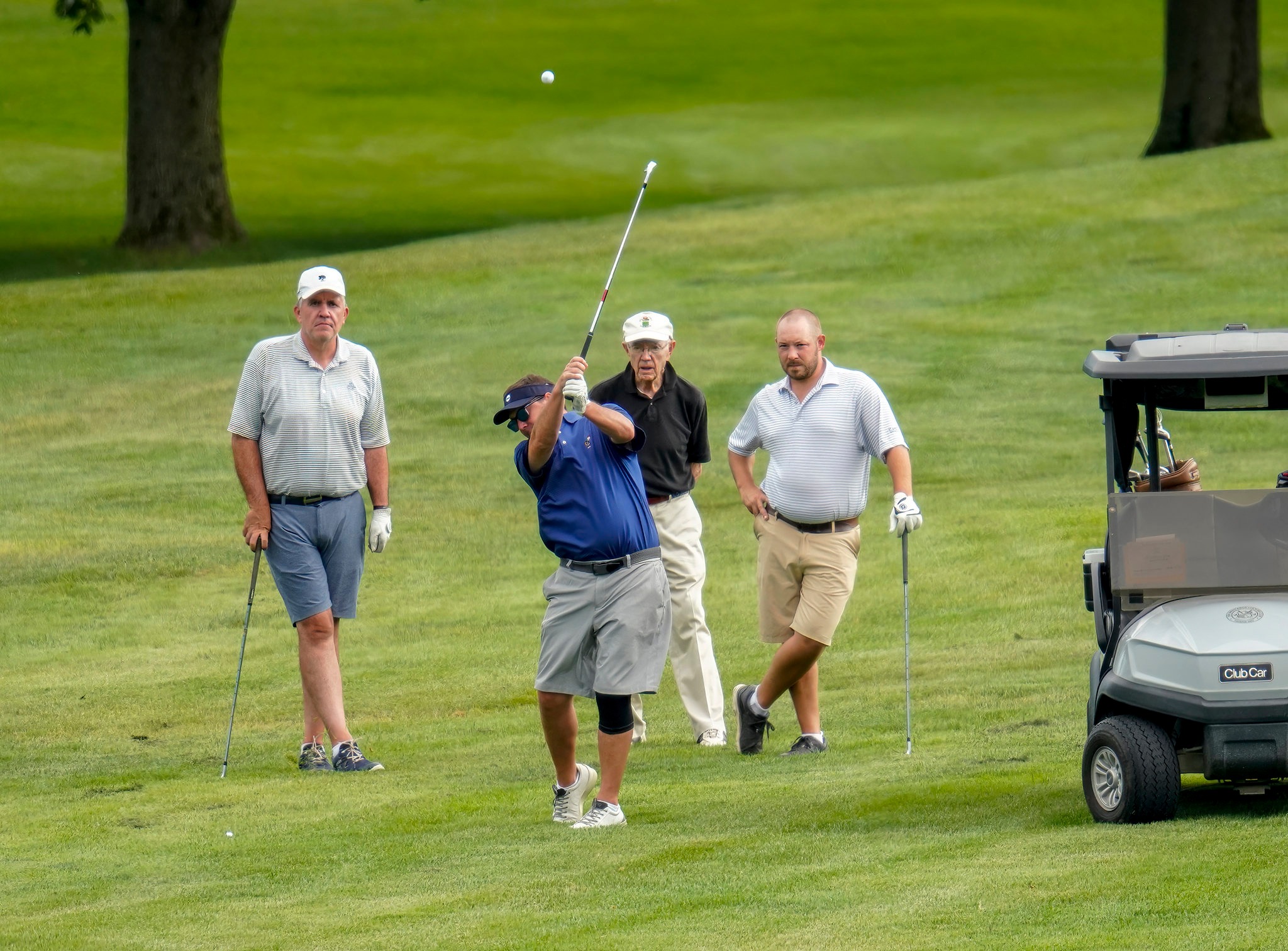 One man takes shot with golf club, while three male companions watch.